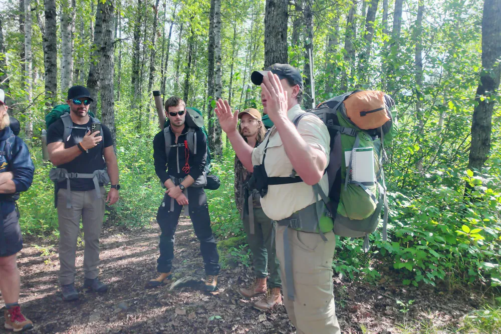 The group standing with packs in the forest listening to their instructor