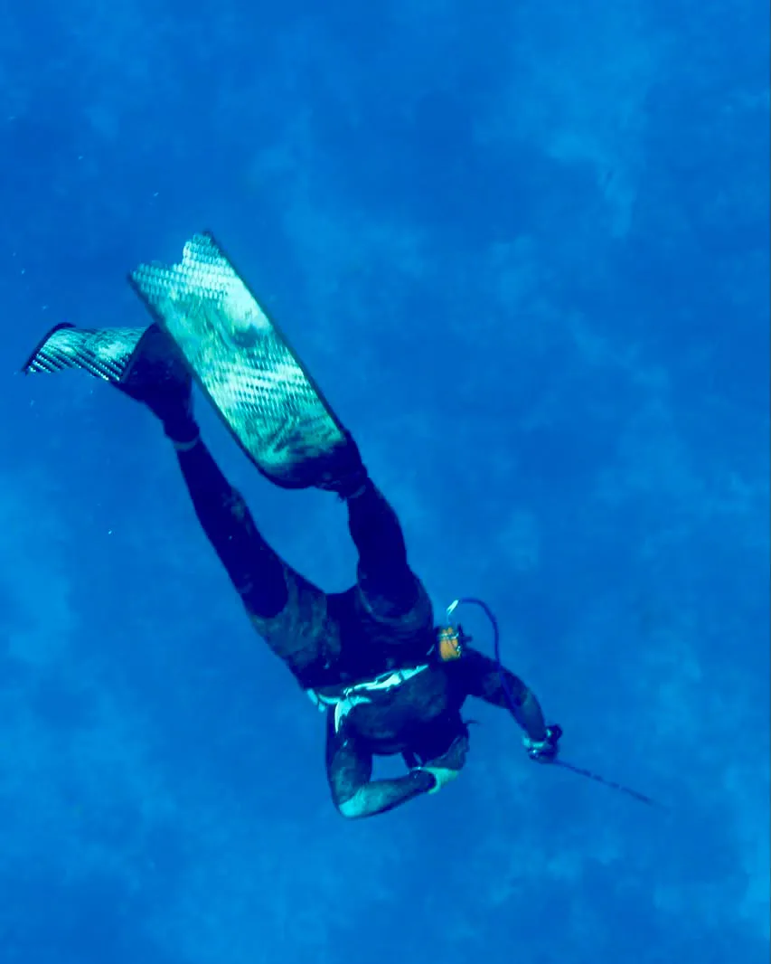 A spearfisher upside down mid descent into deep blue water, fins together and gun in hand
